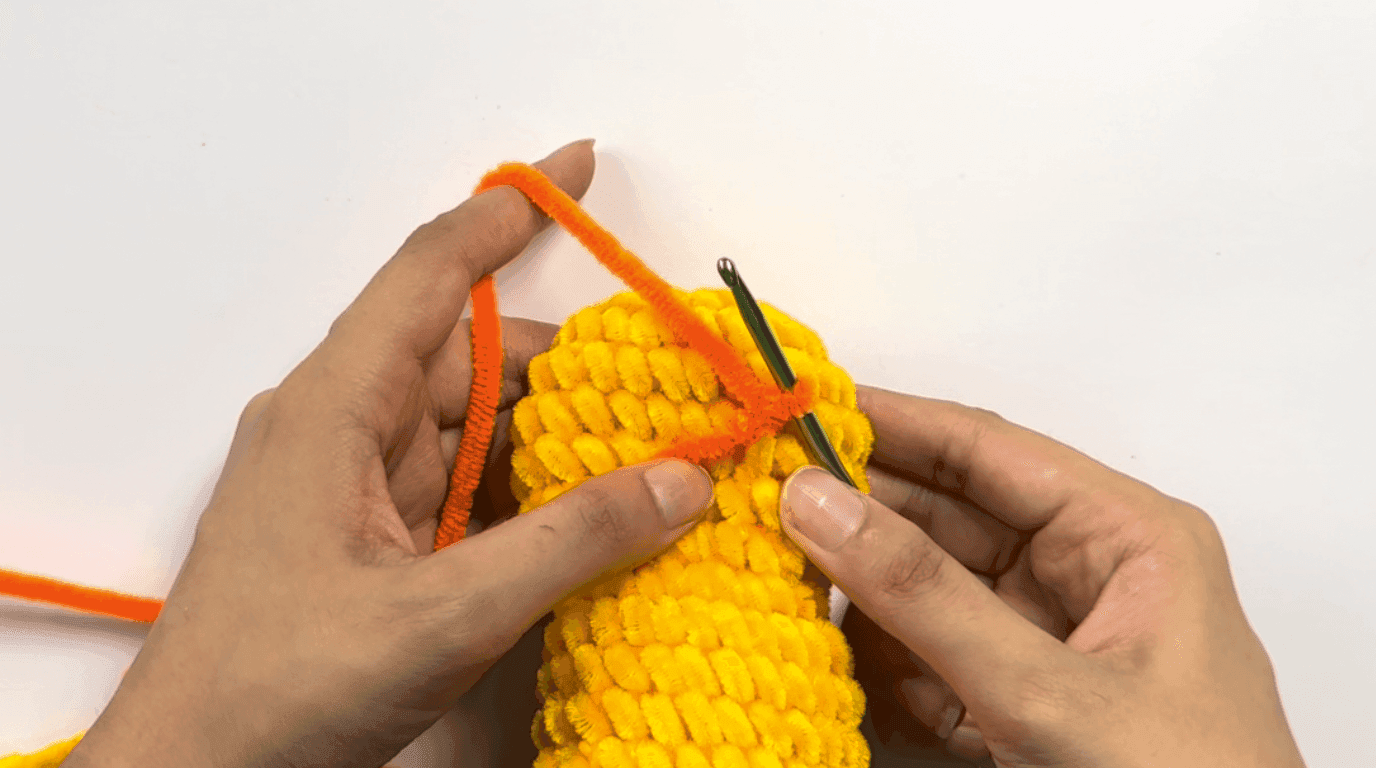 Hands crocheting the first row of the orange beak directly onto the yellow duck body, showing the beak attachment point at the center front.