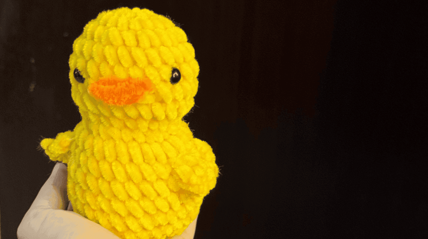 Finished crochet duck pattern held in hand, showing the completed yellow velvet duck with orange beak, black safety eyes, and wings, photographed against a dark background.