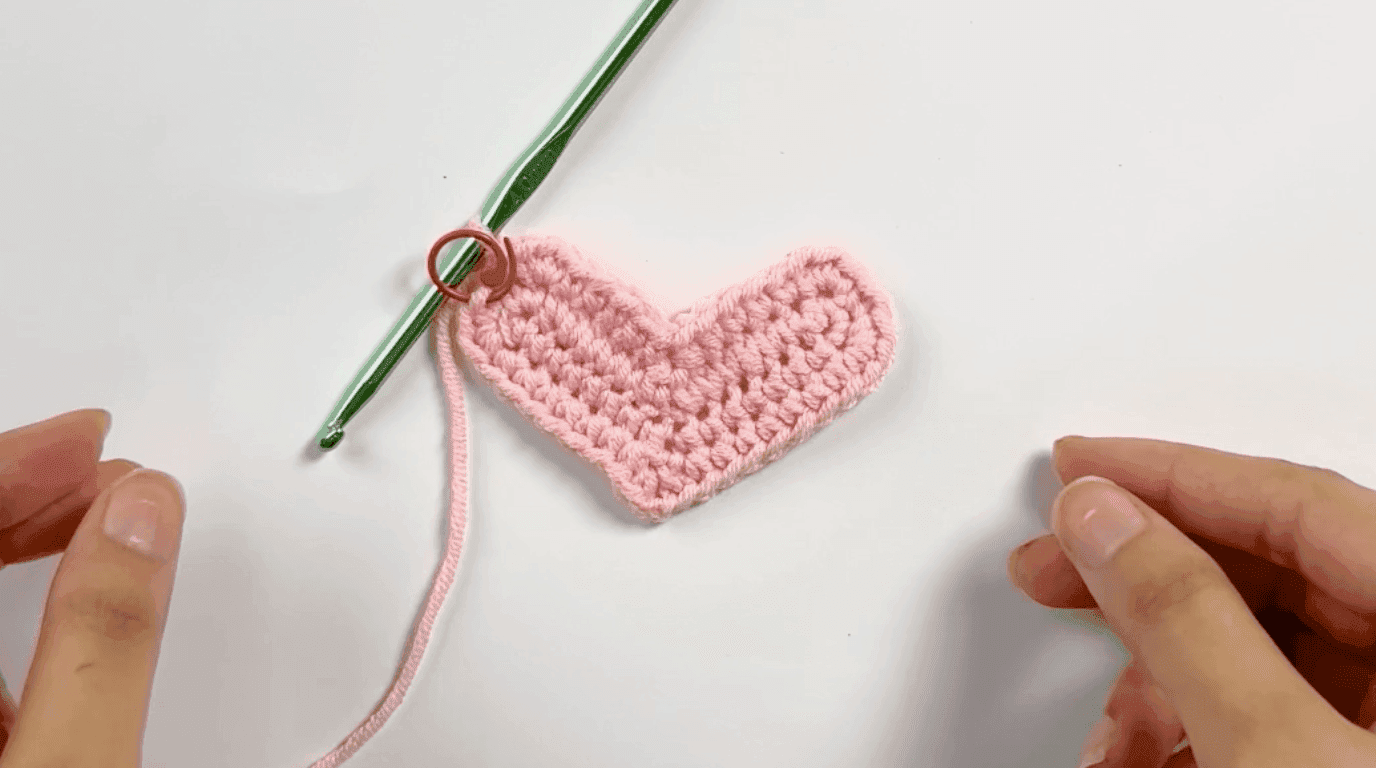 Pink crochet heart base showing progression after Row 2, with hook inserted showing the 3-single-crochet increase technique creating the rounded lobes, white background