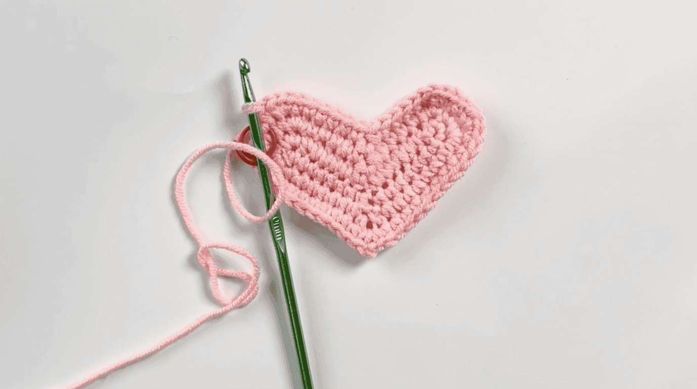 Medium-sized pink crochet heart base after Row 3, showing developing heart lobes and overall silhouette with green hook and working yarn visible, white background