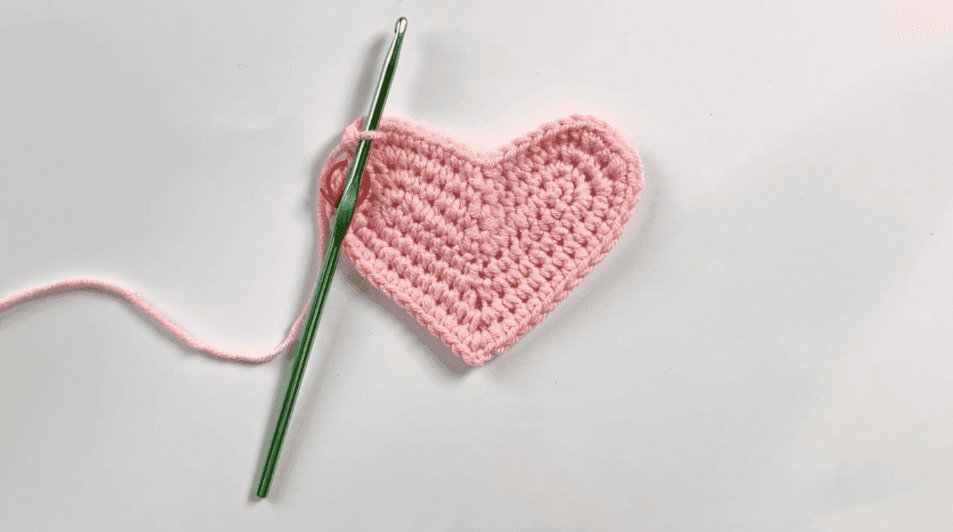 Nearly complete pink crochet heart base after Row 4, showing fully defined two lobes and bottom point with yarn tail, green hook positioned ready to continue, white background