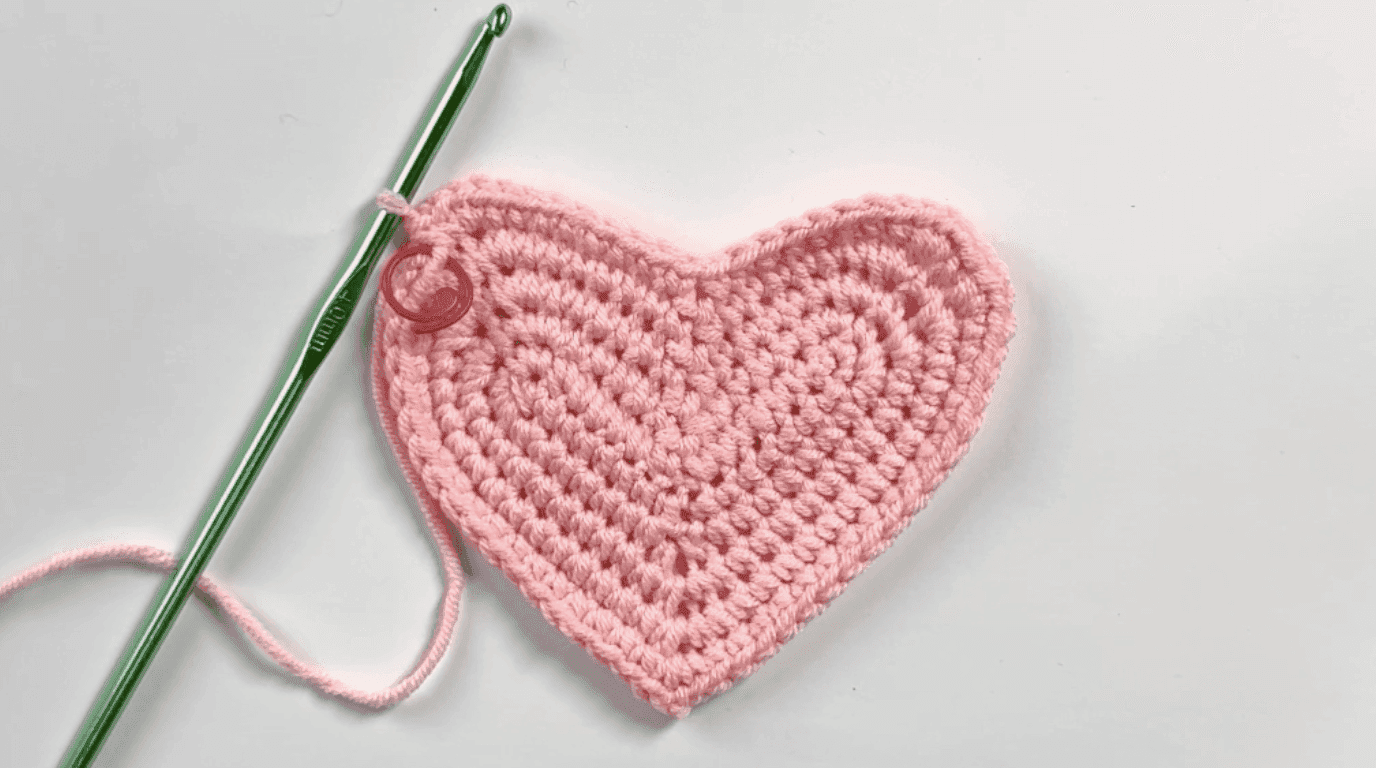 Finished flat heart base after Row 5 with 64 stitches, showing crisp lobes and center indentation with green hook and pink yarn tail, white background