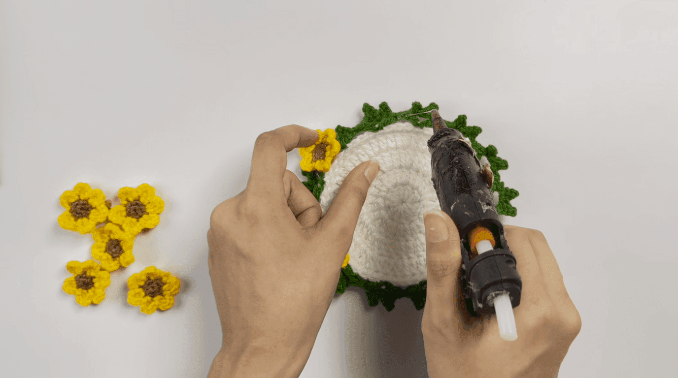 Crochet sunflower pot assembly: hands applying hot glue to flower back with completed coaster and pot nearby on neutral background