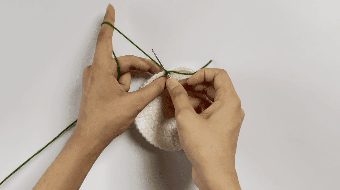 Crochet sunflower coaster leaves Round 1: hands attaching forest green yarn to back loops of coaster edge, showing chain-6 and double crochet foundation technique